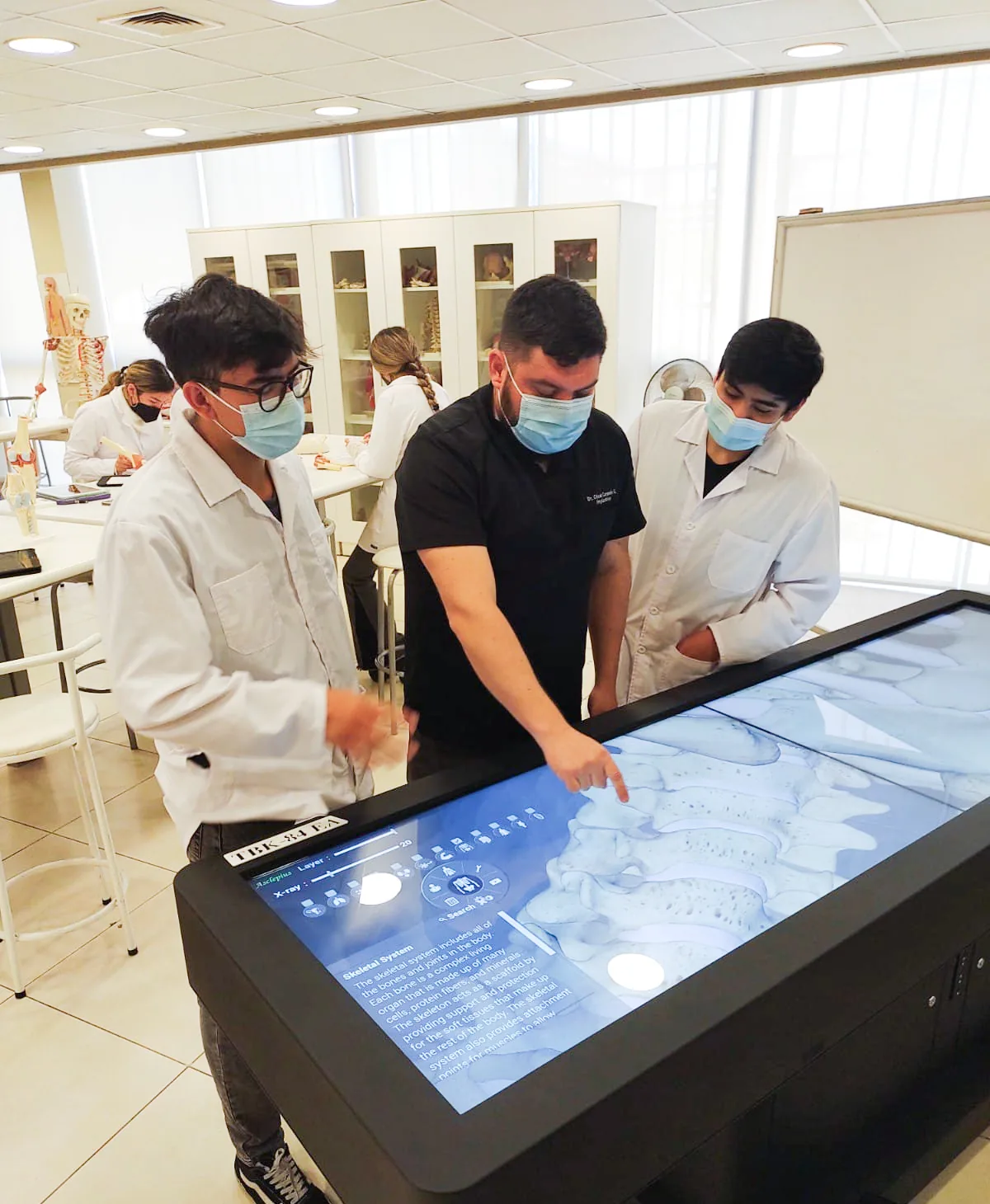 Two students in white coats and an instructor gathered around an upright
                  Asclepius table in an anatomy classroom, examining an X-ray view of the
                  spine on its screen while classmates work at desks behind them.