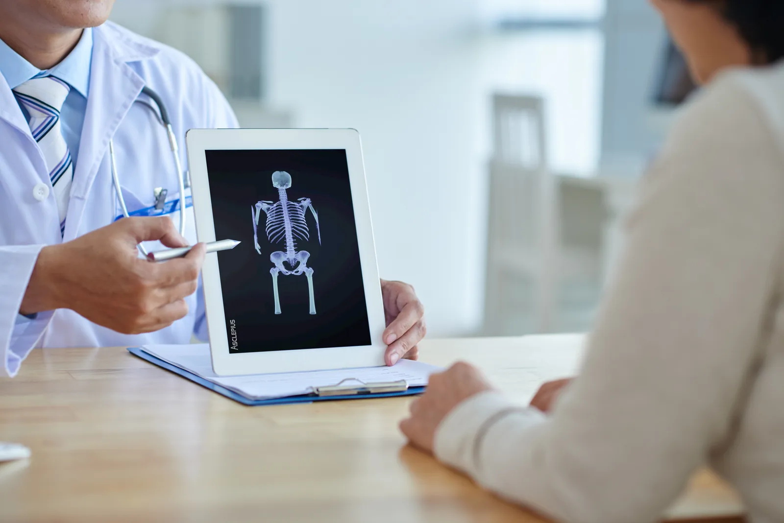 A physician in a white coat holding up a tablet showing an Asclepius skeletal
                  model, pointing at it with a stylus while a seated patient looks on.