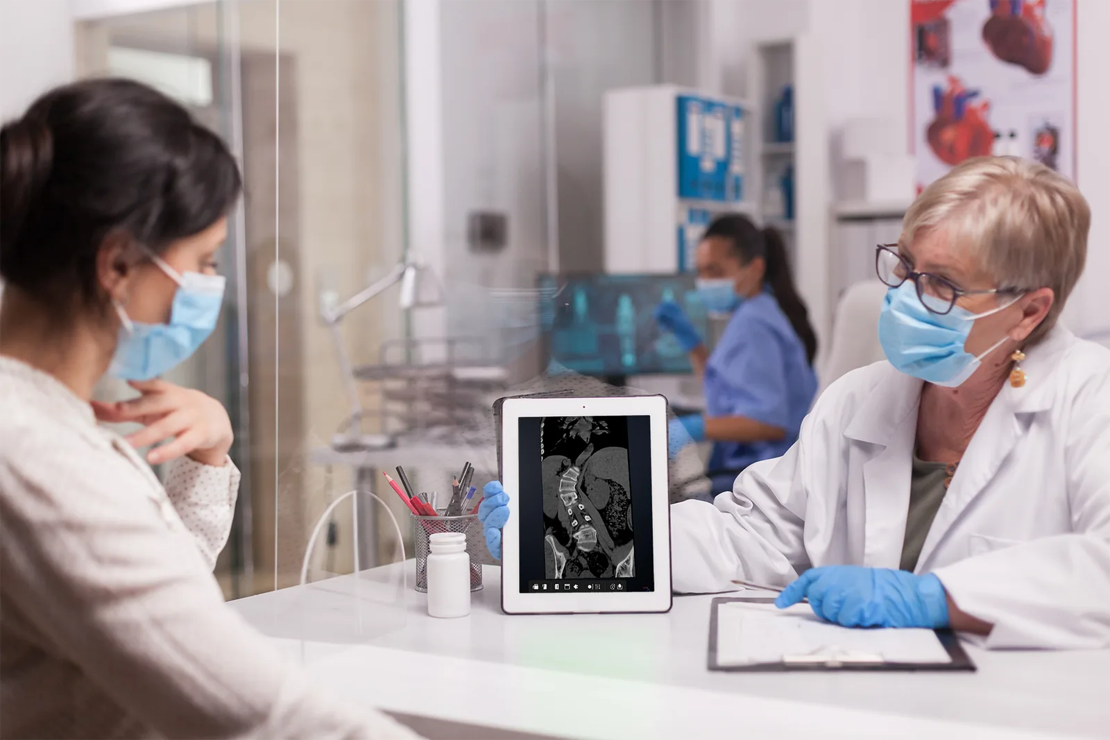 A clinician in a consulting room turning a tablet towards a patient to
                    show a spinal CT scan on its screen.