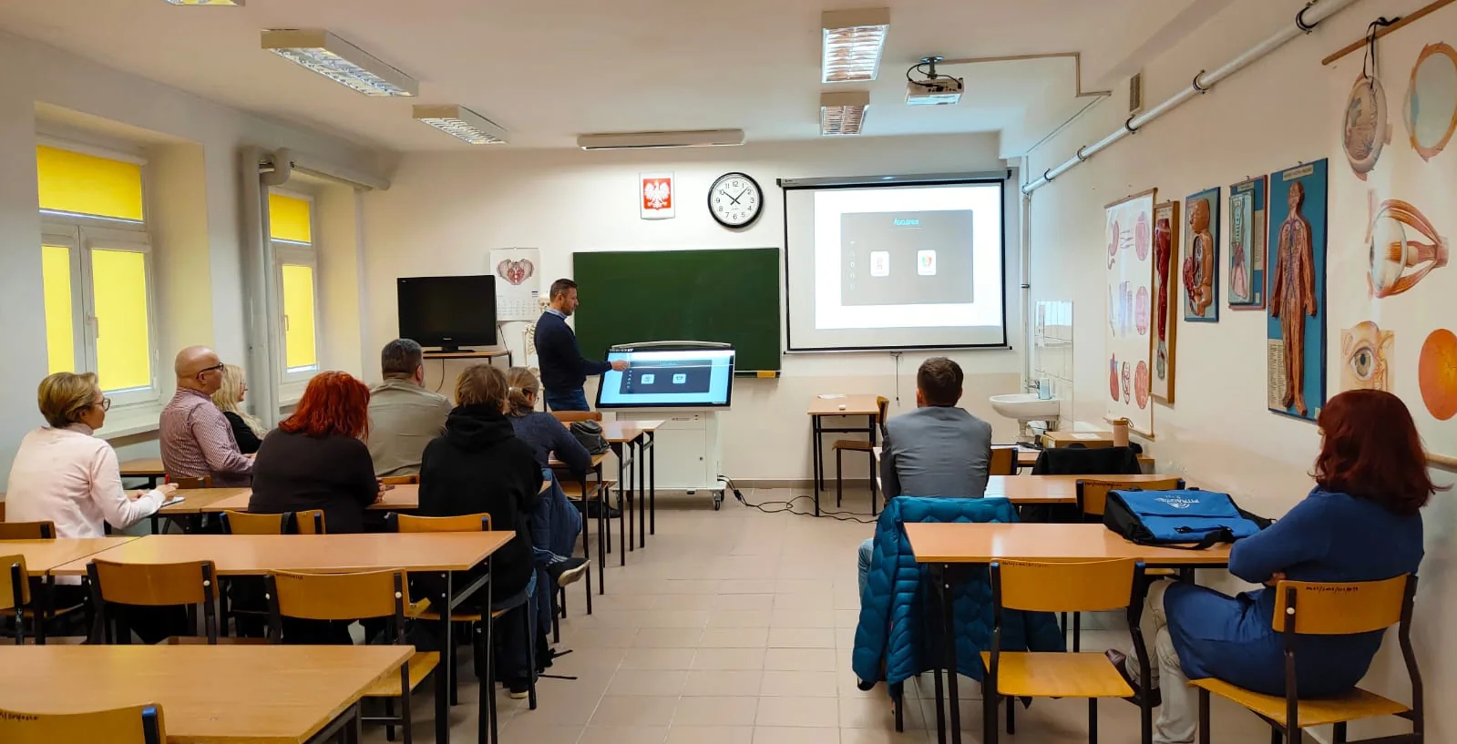 A seminar room of seated adults watching an instructor operate an Asclepius
                  table, the table's screen mirrored onto a projector screen beside the
                  blackboard and anatomical charts on the wall.