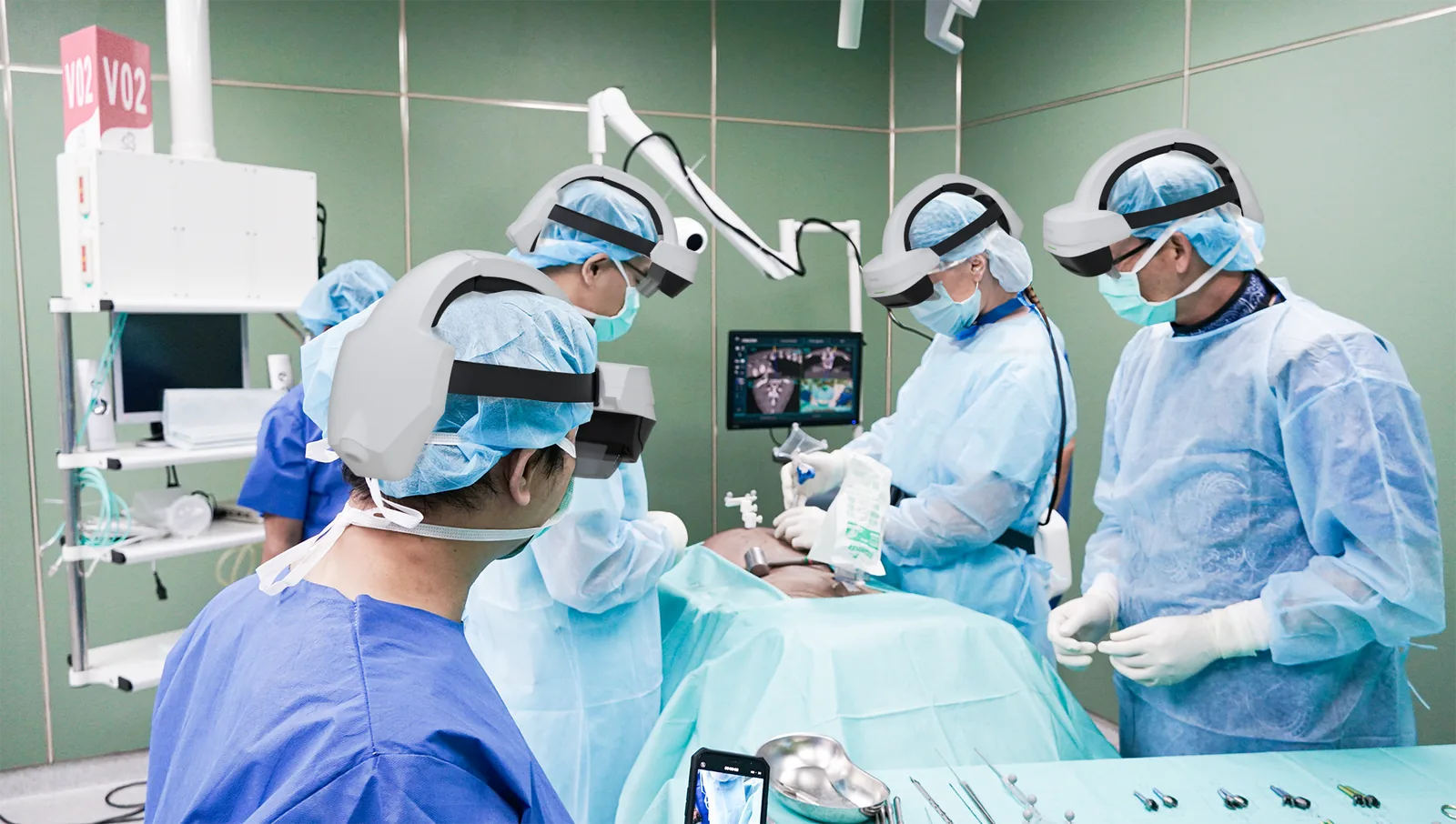 Four members of a surgical team in an operating theatre, each wearing a
                  Caduceus S headset over their surgical cap, gathered around a draped patient
                  with imaging displayed on a monitor behind them.