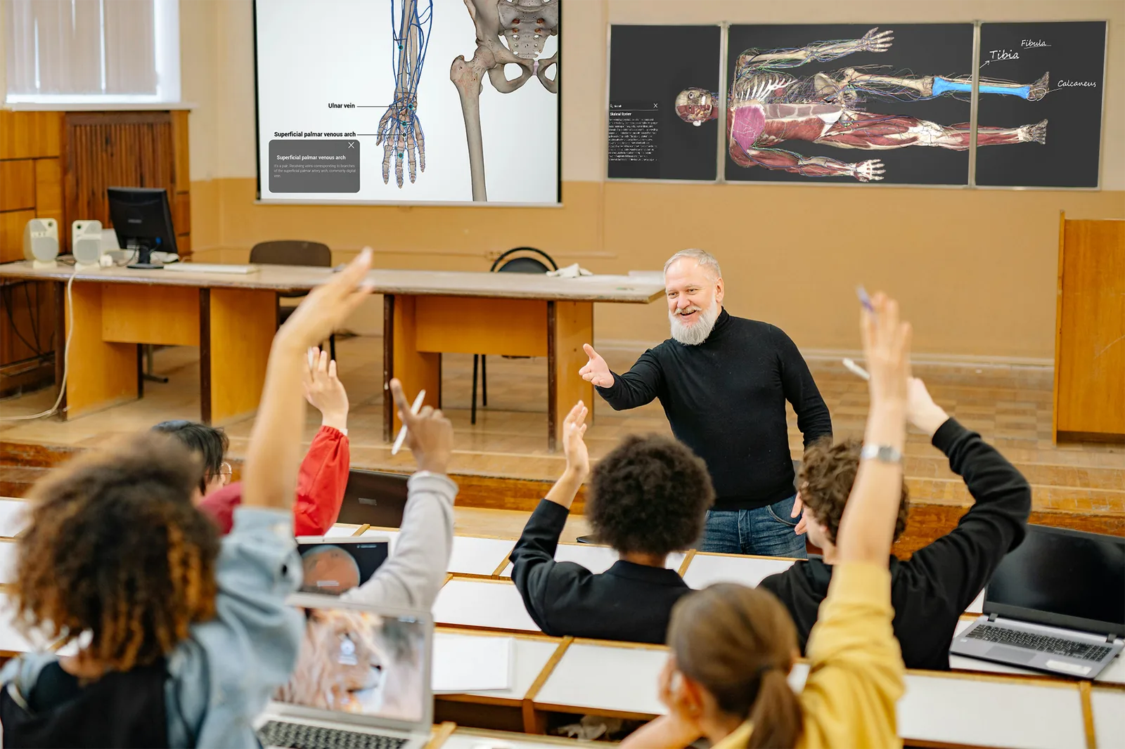 A lecture room of students with their hands raised, facing a projected
                    Asclepius vascular diagram and a wall panel showing a full-body dissection
                    view.