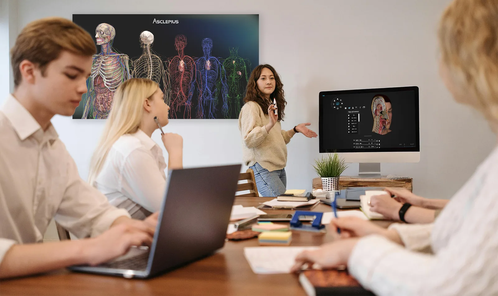 Students working at laptops around a table while a classmate presents an
                    Asclepius body-systems display on the wall screen behind her.