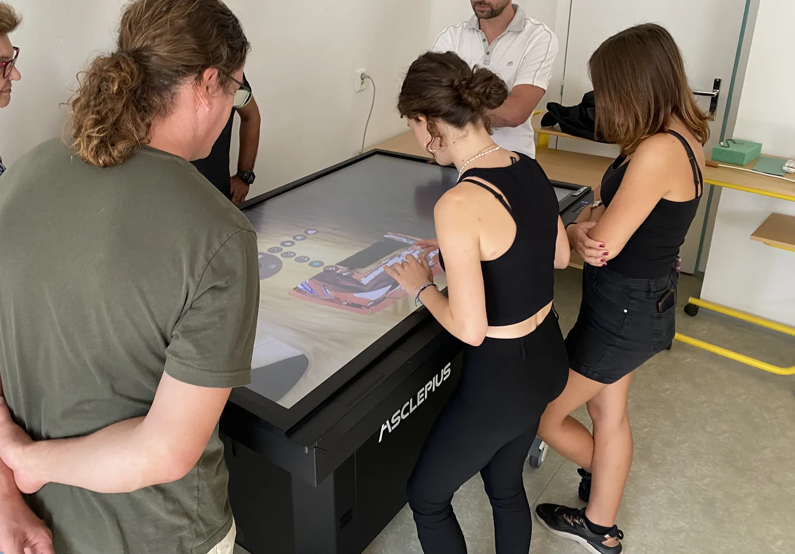 A group of students leaning over an Asclepius table together, one of them
                    manipulating an anatomical model on the touchscreen.