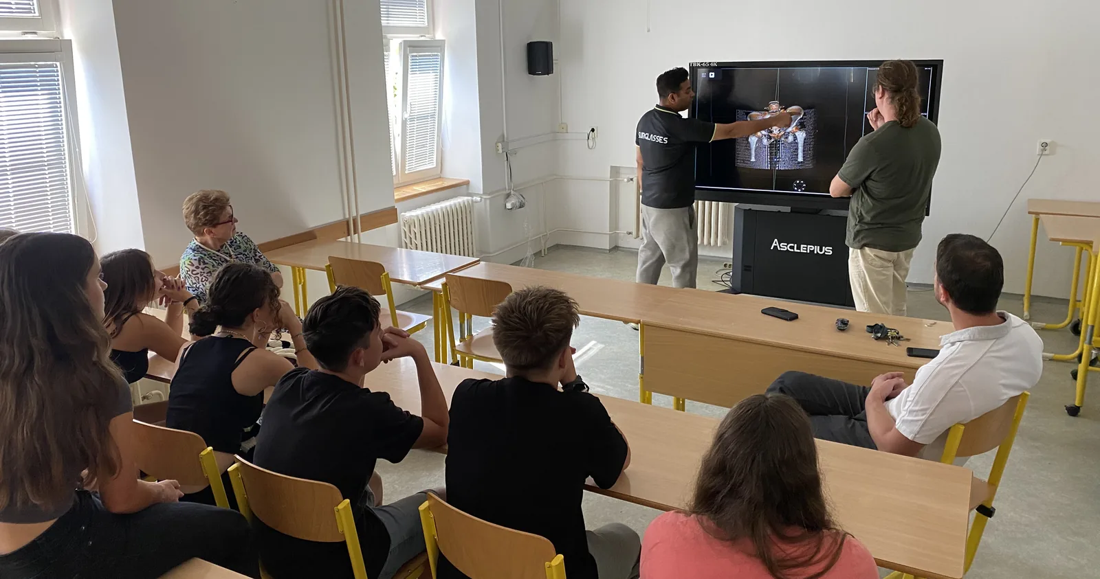 A class seated in a bright teaching room watching two instructors work at an upright Asclepius dissection table
