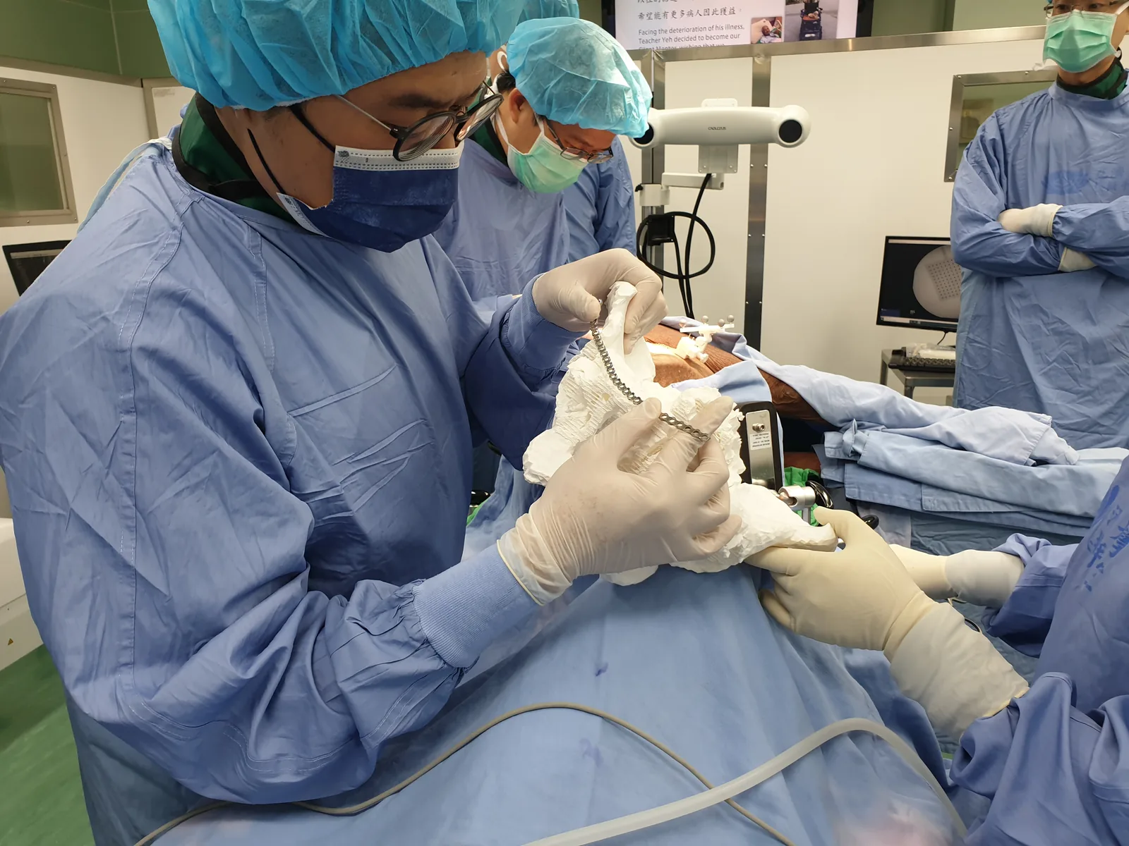 In an operating room, a gowned and gloved surgeon holds a white 3D-printed
                  pelvis in one hand and a metal fixation plate against it in the other,
                  contouring the plate to the bone over the draped patient while the rest of
                  the team looks on.