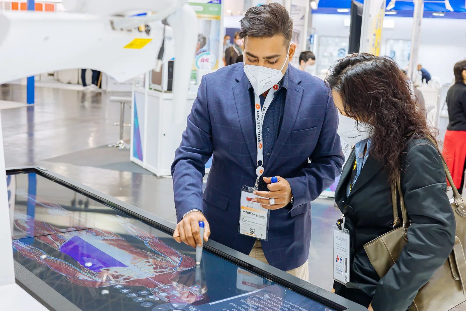 Two visitors at the Surglasses stand at the MEDICA trade fair, one of them using a stylus on a life-size anatomy rendering shown on the virtual dissection table