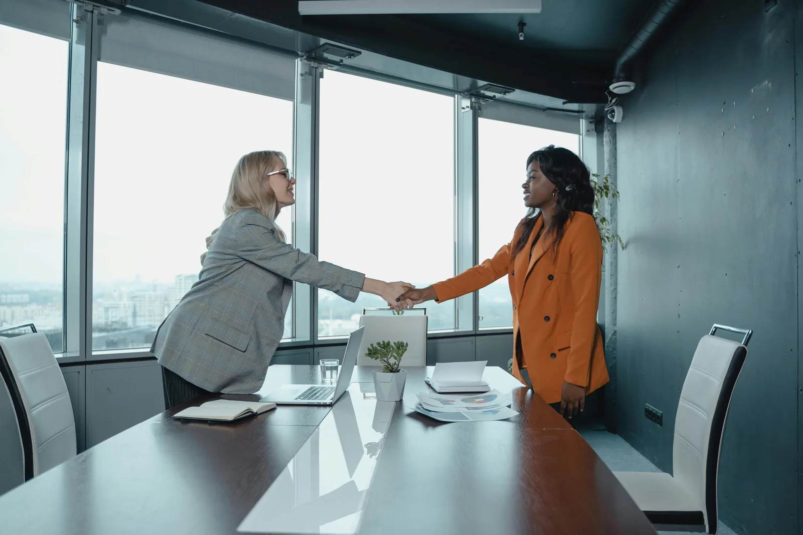 Two people shaking hands across a meeting-room table, with a city skyline through the windows behind them
