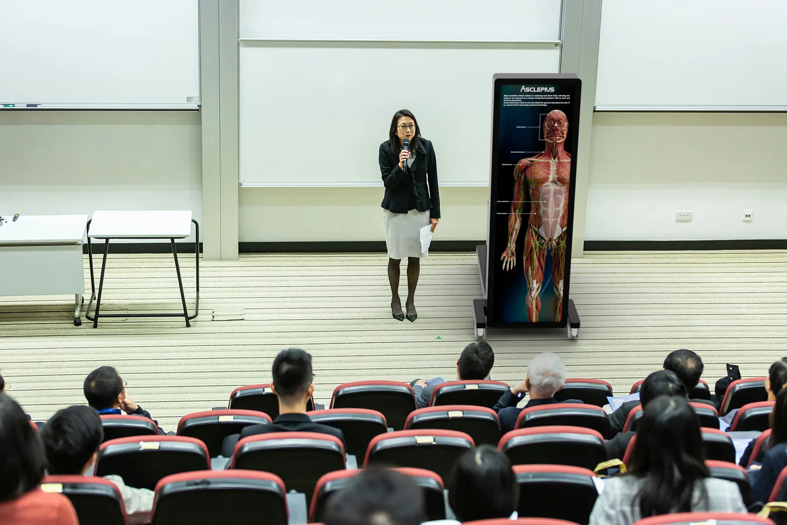 A TBK-84 EA table standing upright on a lecture theatre stage, showing a
                    full-body anatomical model to a full auditorium.
