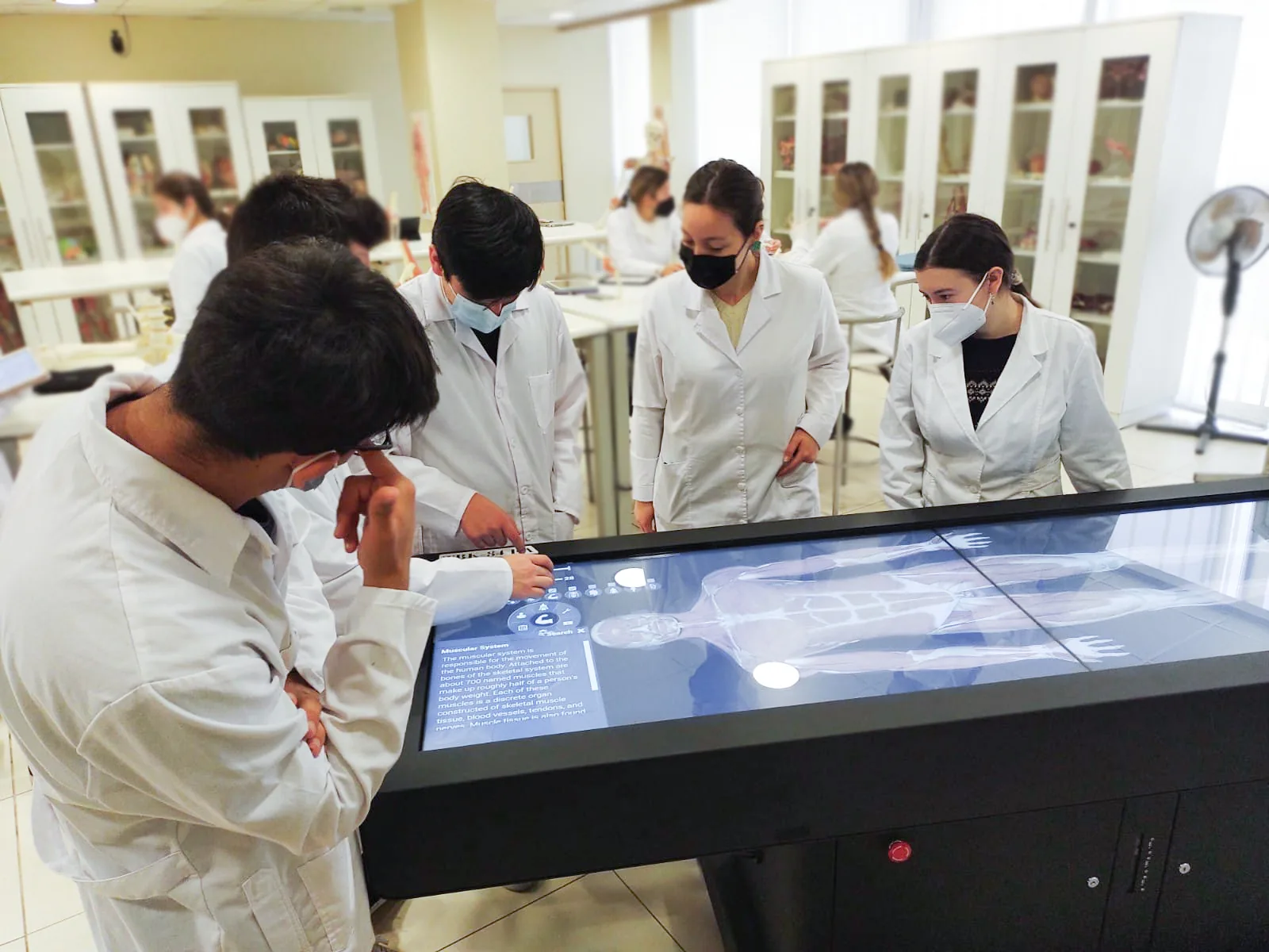 Medical students in white coats working with a life-size anatomy rendering on an Asclepius table in a teaching laboratory